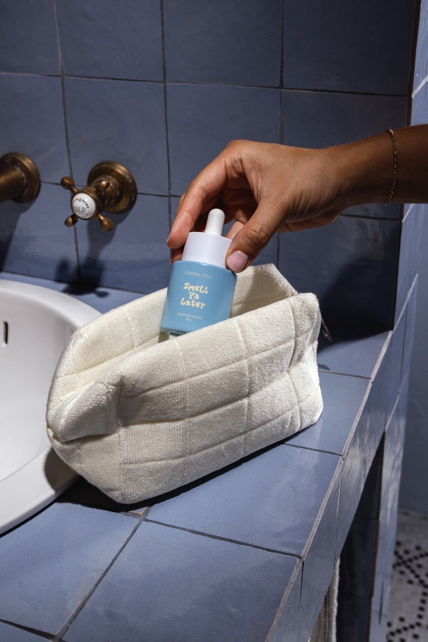 Hand holding a blue bottle with a pump over a textured white bag in a bathroom setting.